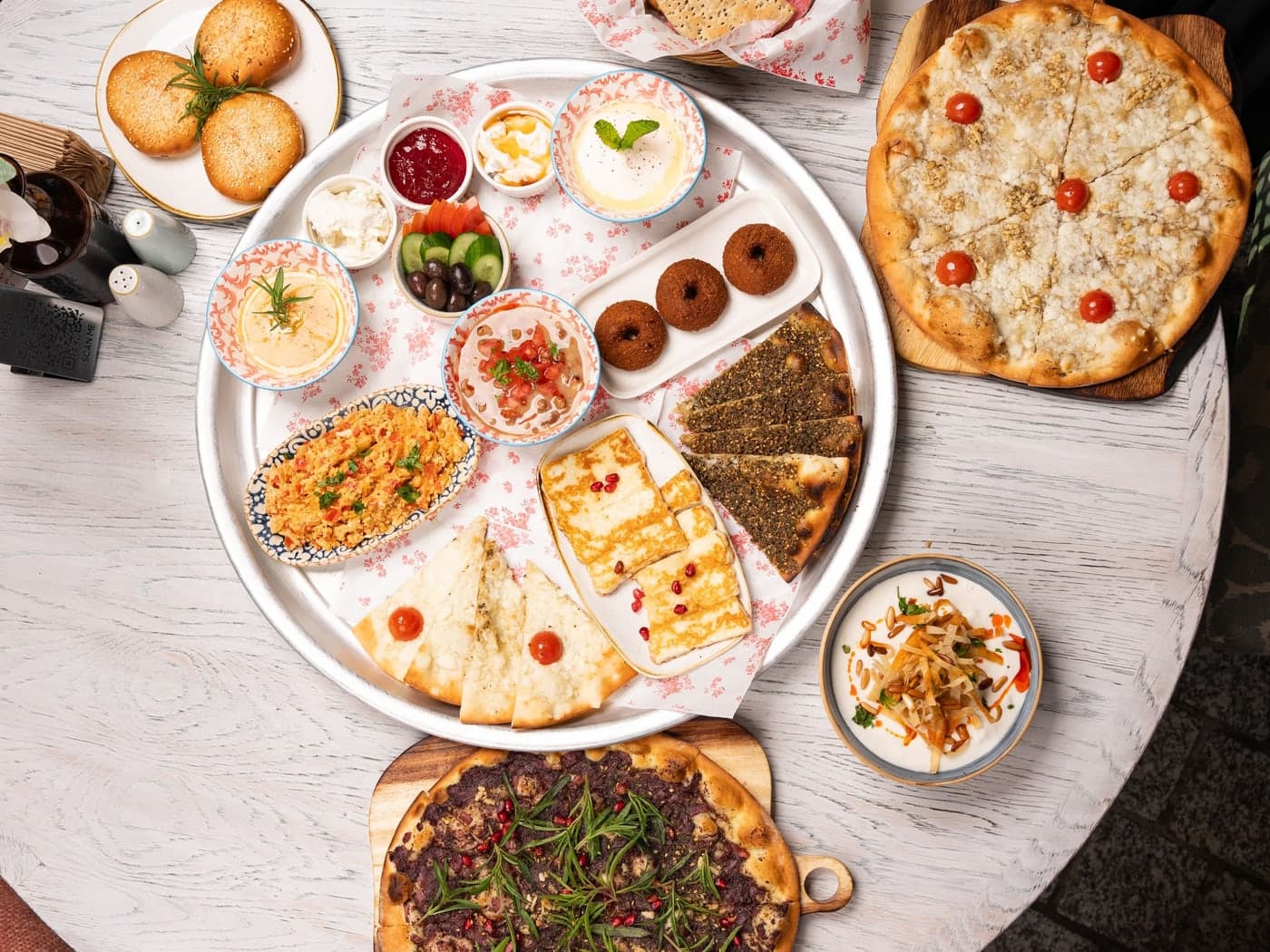 An overhead Levantine mezze spread on a pale wooden table — hummus, labneh, olives, grilled halloumi and za'atar flatbreads