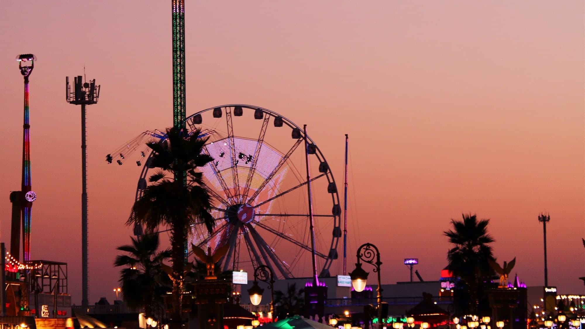 A fairground at sunset in silhouette, a big wheel and a drop tower against a pink and violet sky