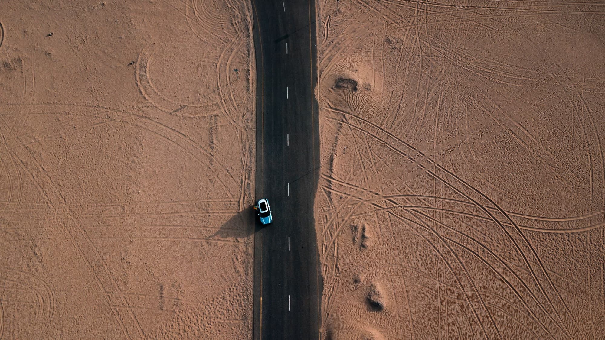 An aerial view of a two-lane road running straight through open desert, a single car on it and tyre tracks fanning across the sand