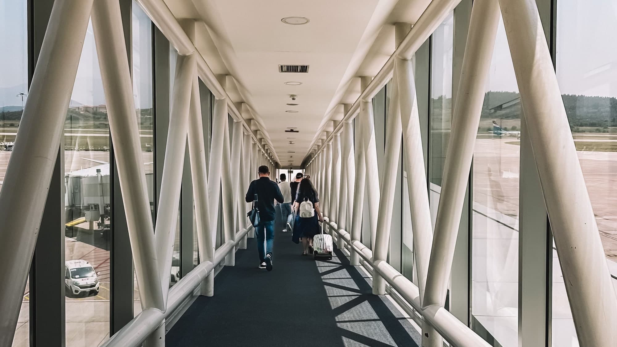 Passengers walking away down a white steel-and-glass jet bridge, the airport apron visible through the glass on both sides