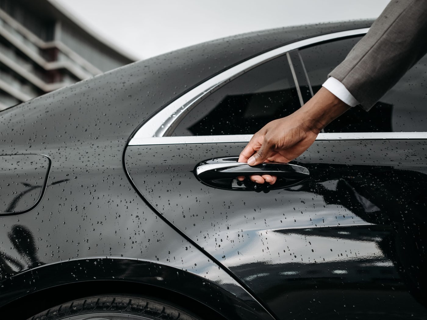 A suited arm reaching for the rear door handle of a black car beaded with rain
