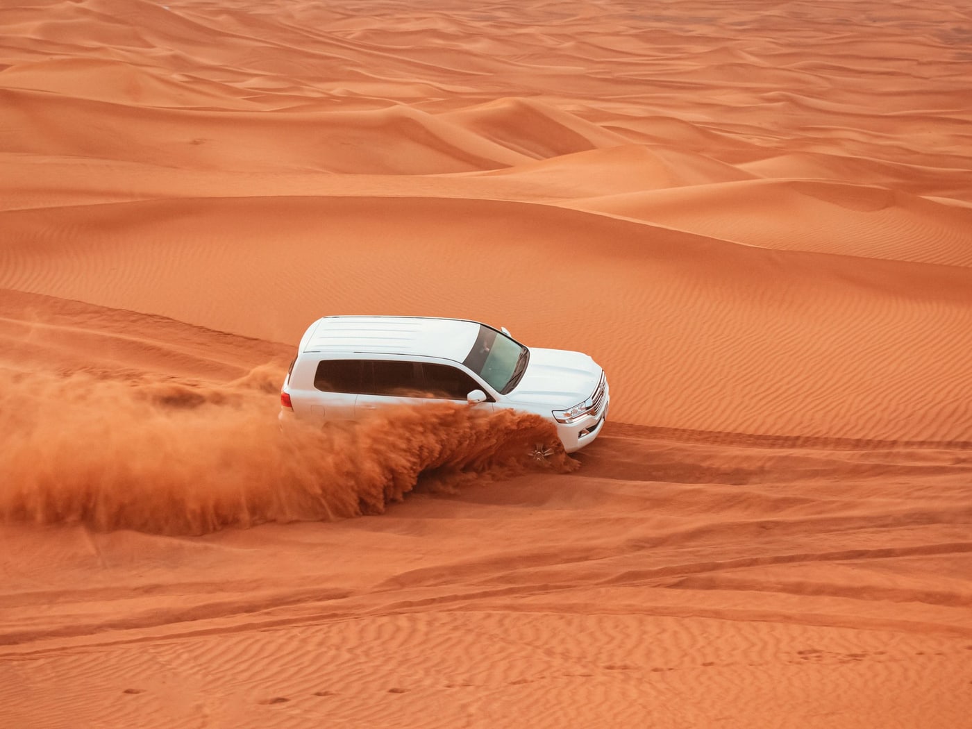 A white four-wheel drive driving across red dunes, throwing a fan of sand behind it