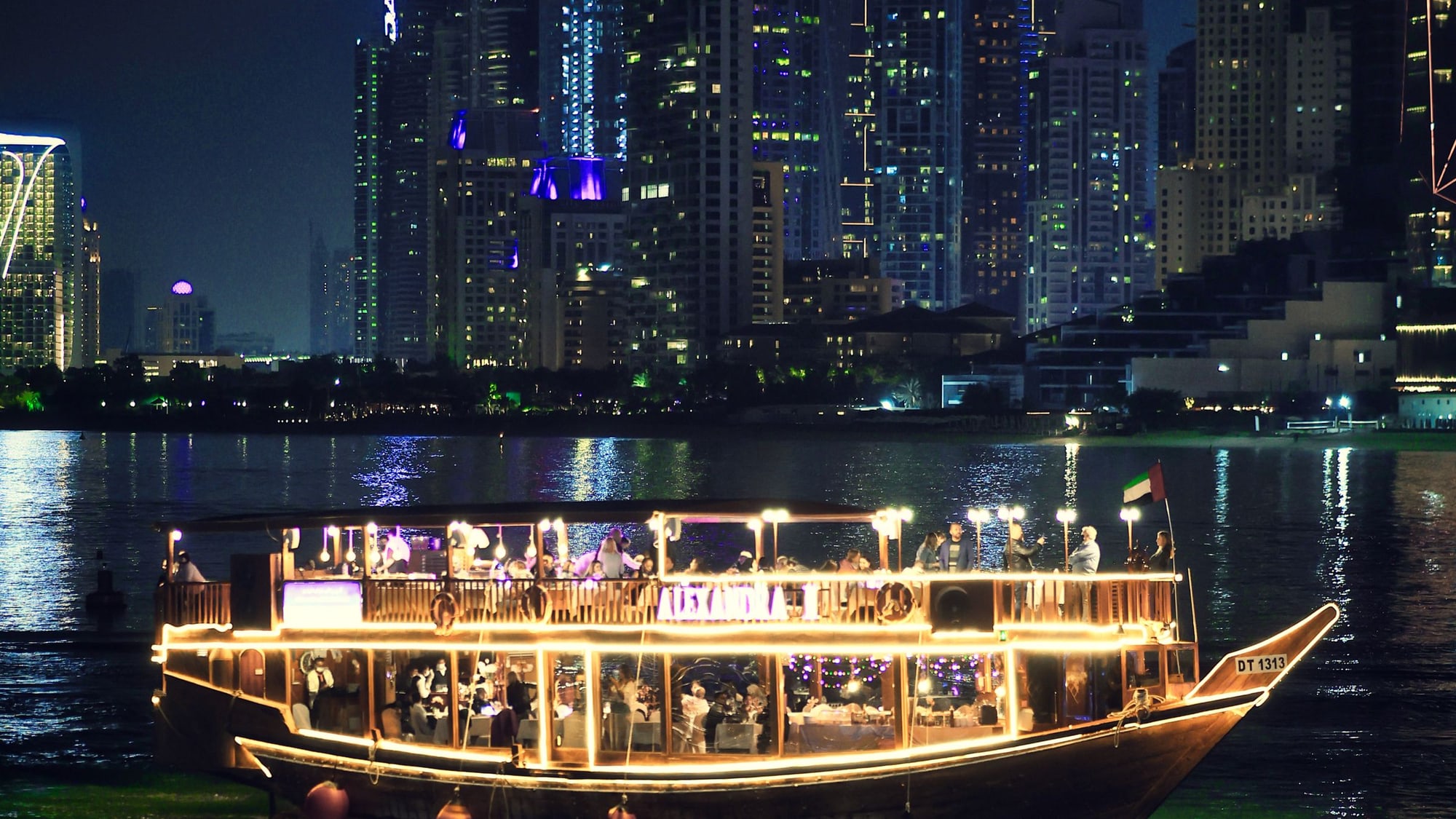 A dhow dinner cruise lit end to end at night on the Dubai Marina water, guests dining on both decks and the Marina towers behind