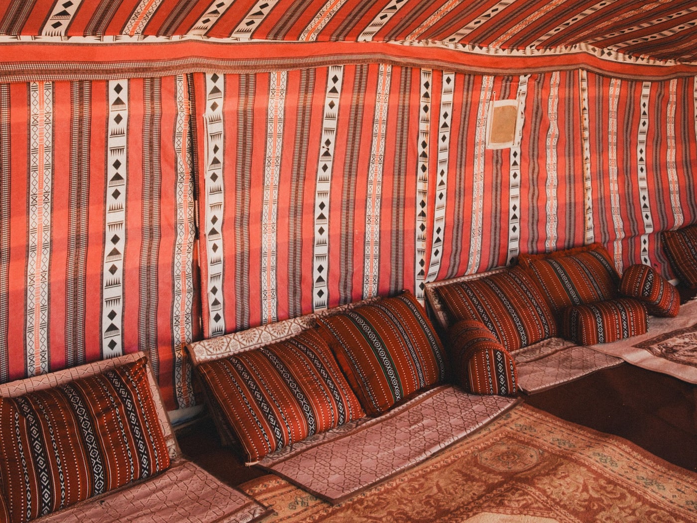 The inside of a Bedouin-style camp tent, red and orange striped panels above patterned floor cushions and carpets