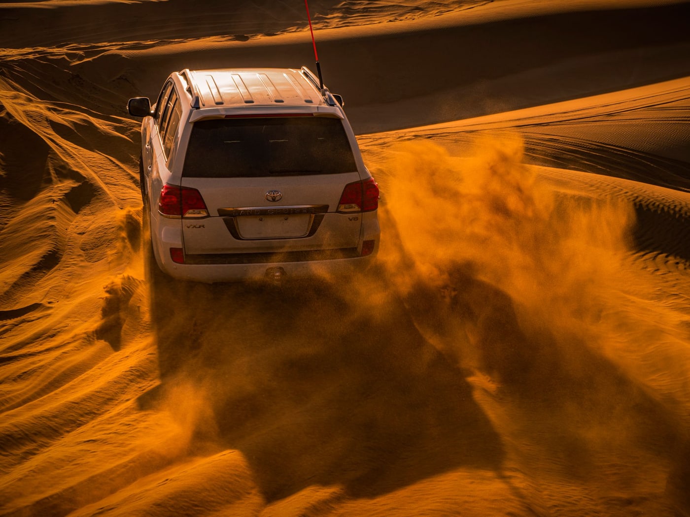 A white four-wheel drive cresting a red dune in warm low light, sand streaming off the ridge behind it