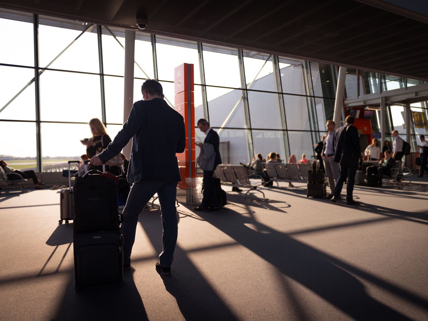 Travellers crossing a sunlit airport terminal with wheeled luggage, long shadows across the floor