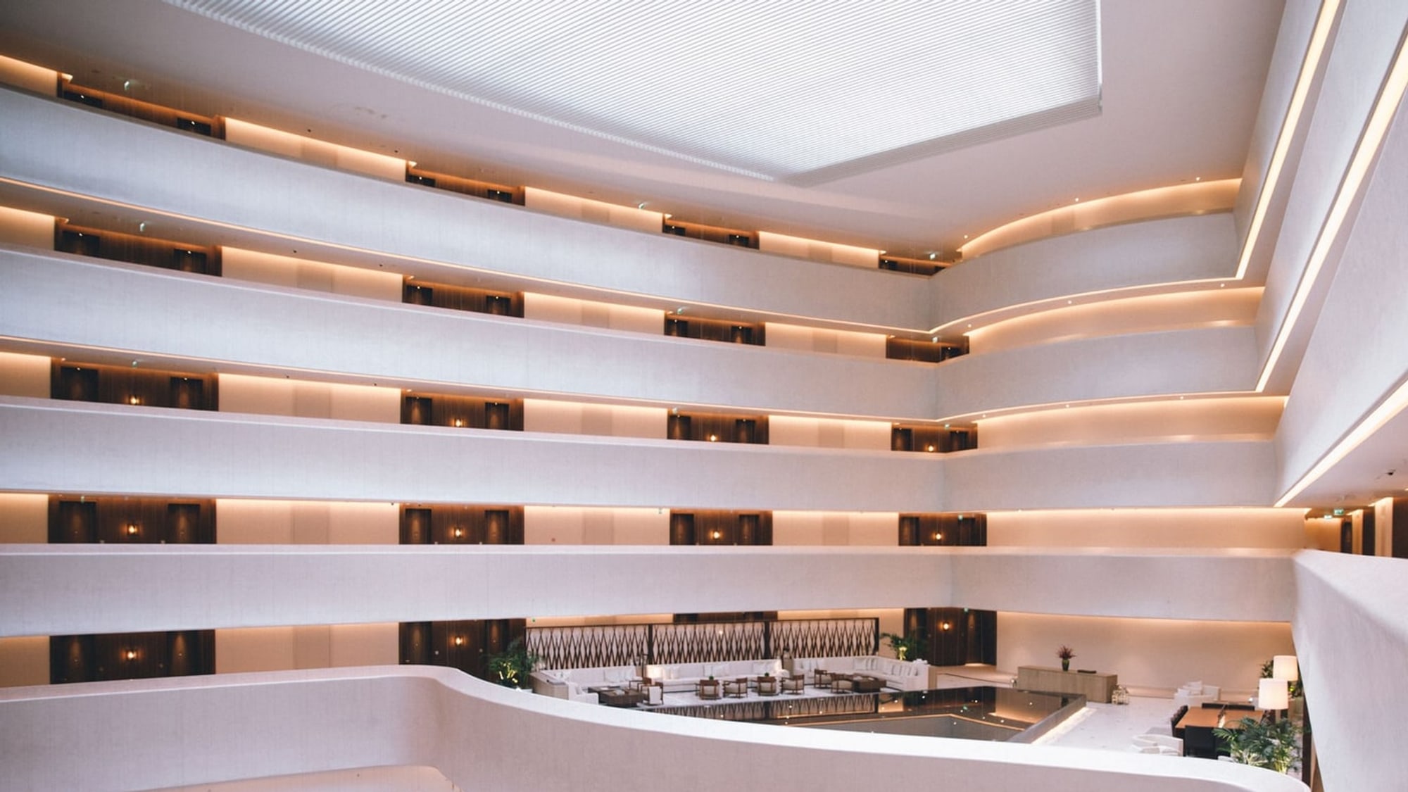 The interior void of a hotel atrium, tiers of curved cream balcony galleries lit by warm cove lighting above a ground-floor lounge
