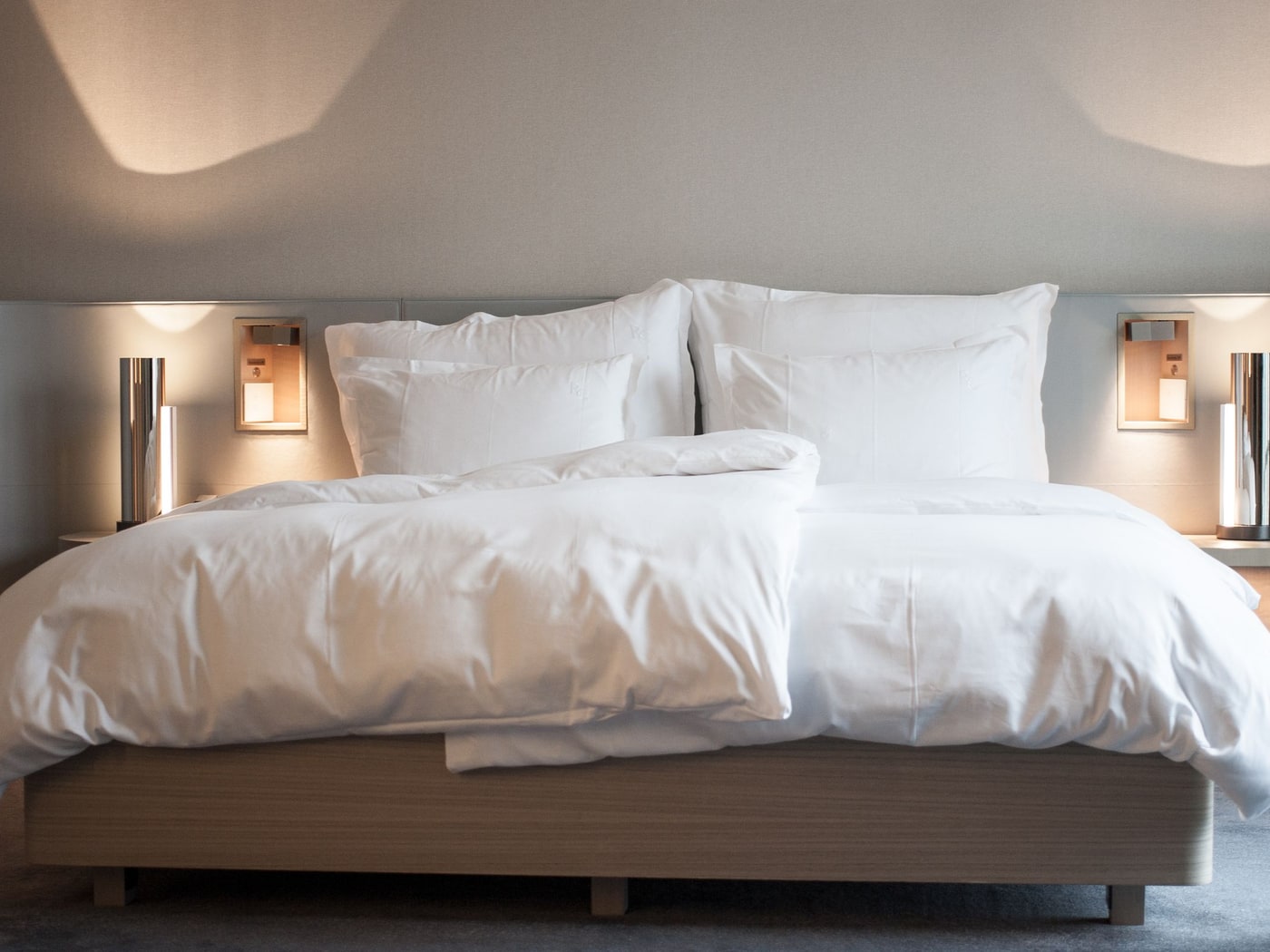 A made-up hotel bed with a white duvet and stacked pillows against a low upholstered headboard, warm sconce light either side