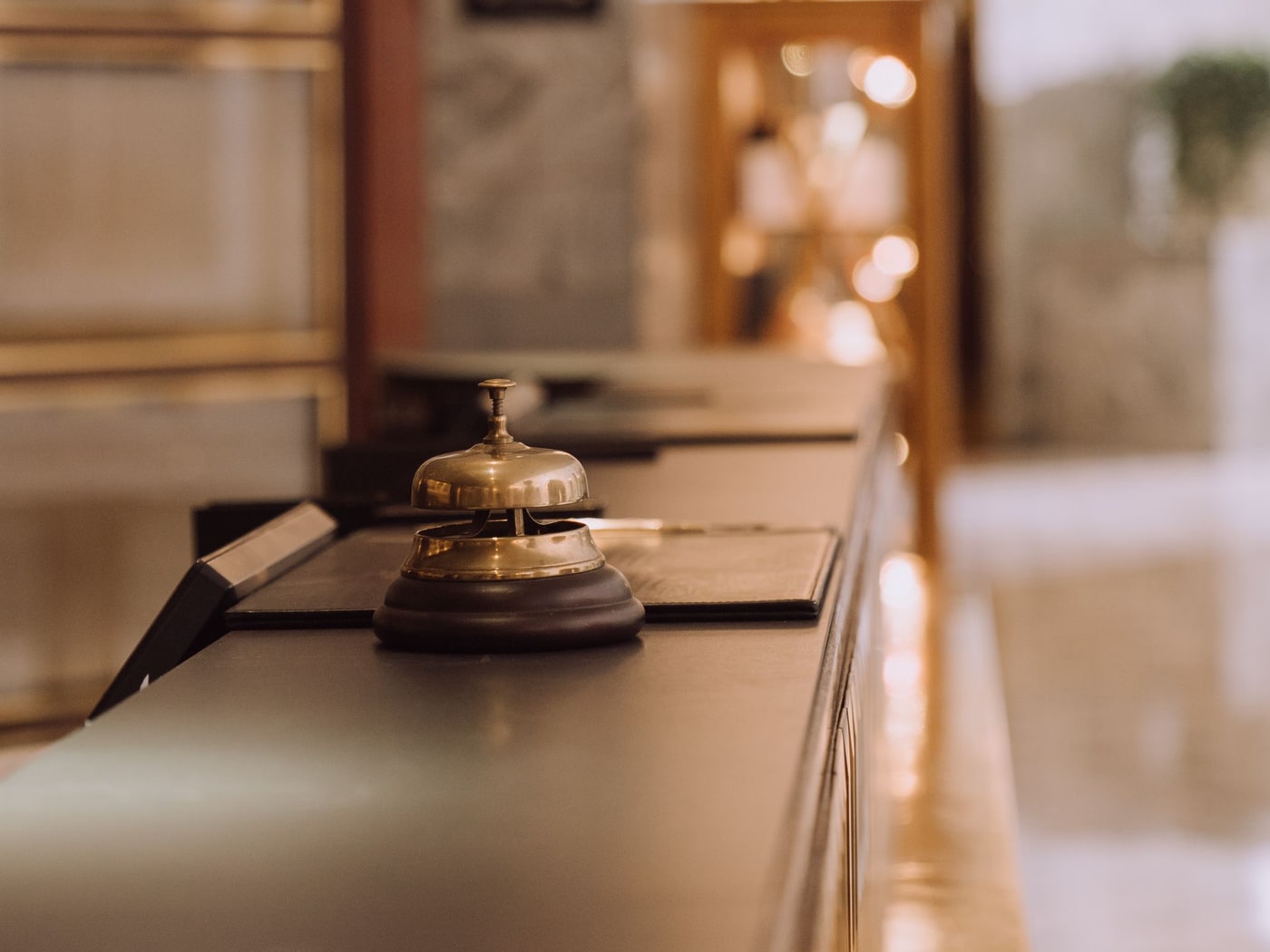 A brass service bell on a turned wooden base on a reception counter, the lobby behind it dissolved into warm bokeh