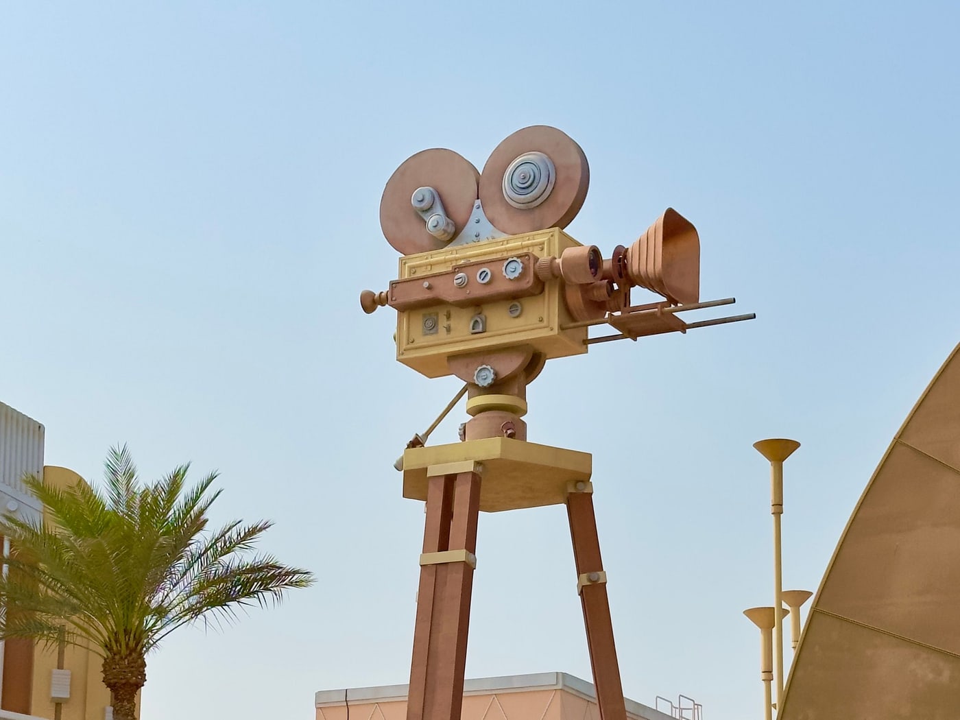 An oversized gold film-camera sculpture on a plinth beside palms at a park entrance