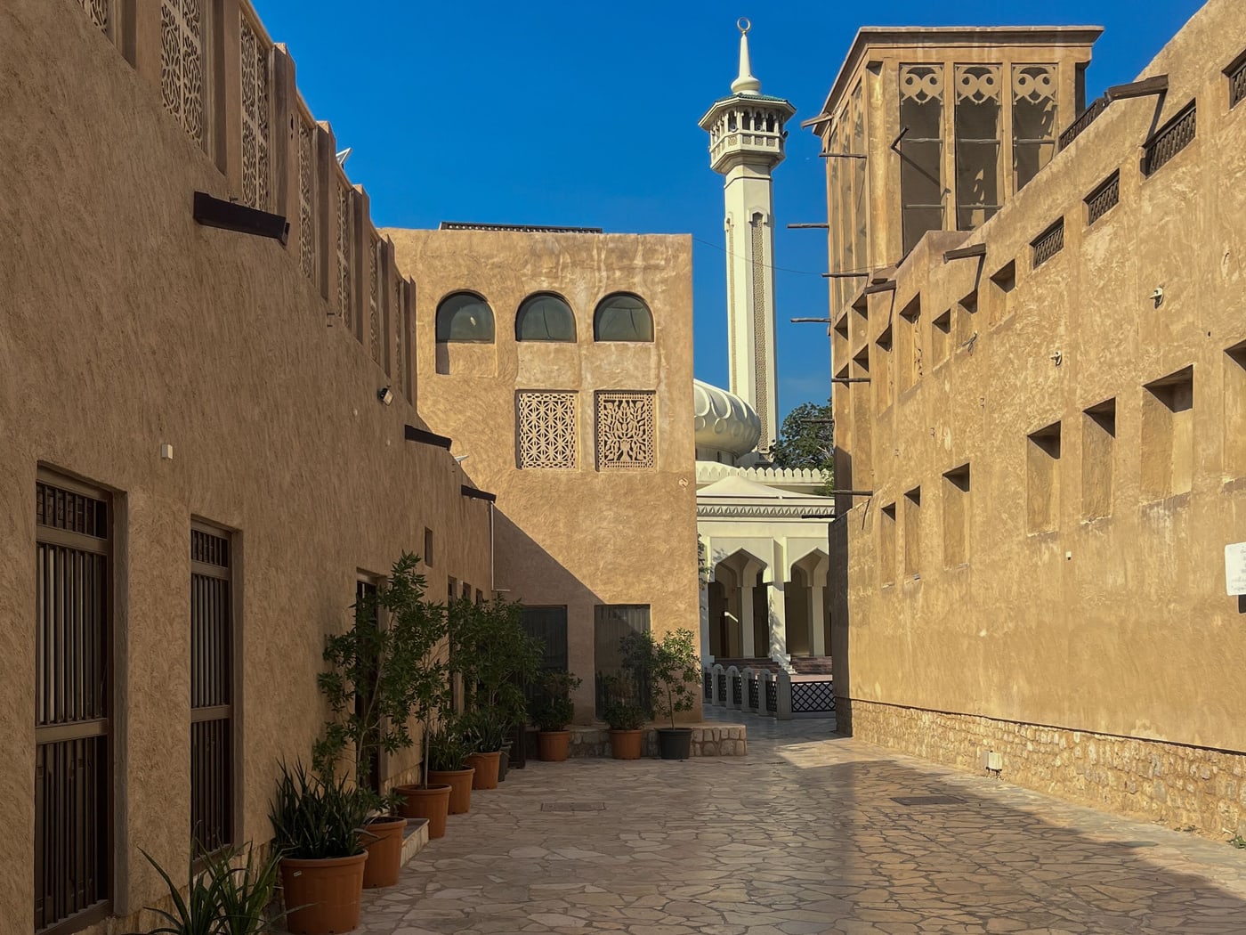 A sunlit lane in an old quarter, sand-coloured walls and wind towers either side and a minaret at the end