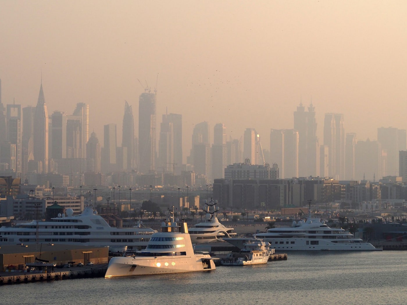The Dubai skyline seen across a marina in afternoon haze, moored yachts along the near shore