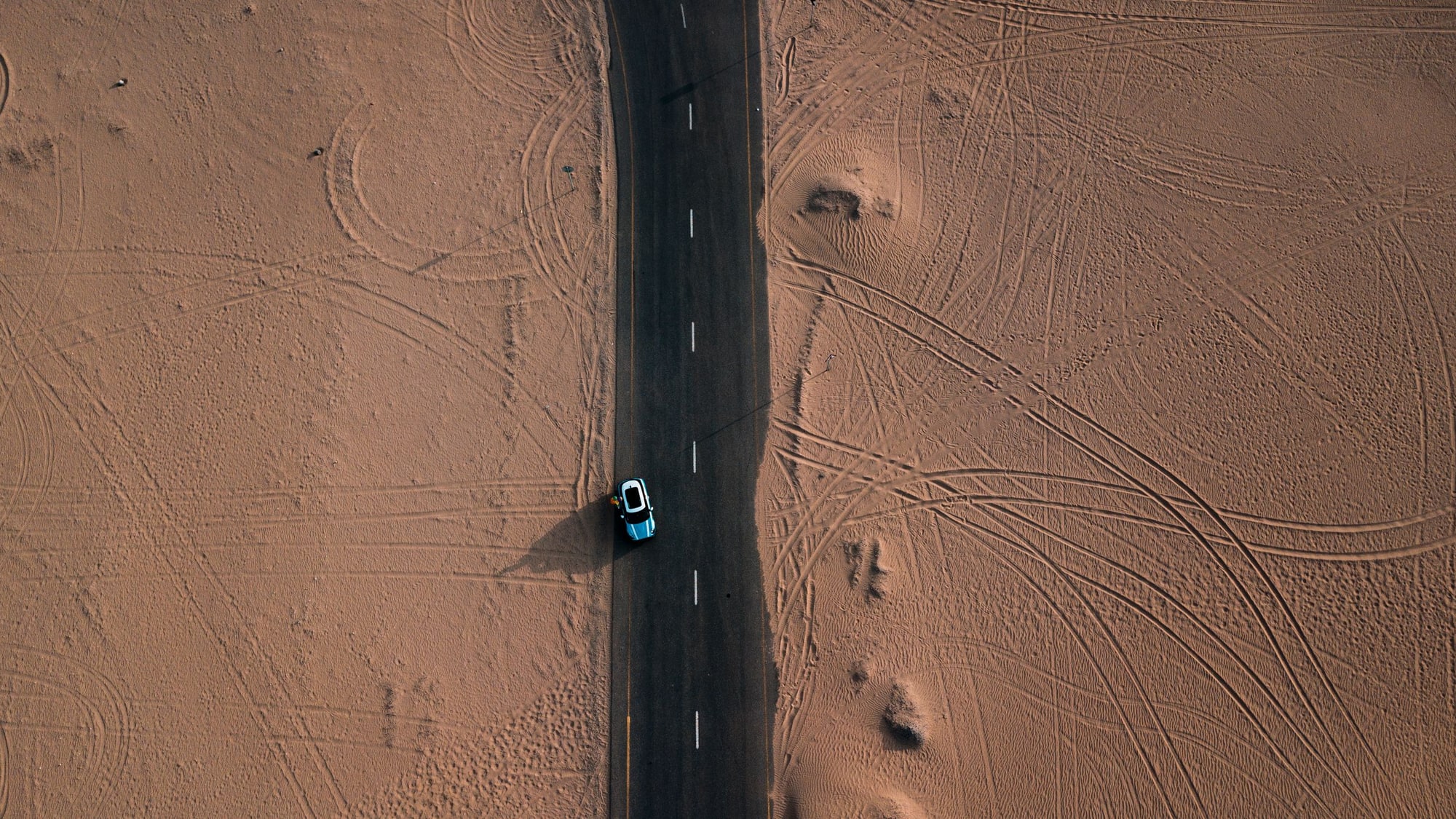 An aerial view of a two-lane road running straight through open desert, a single car on it and tyre tracks fanning across the sand