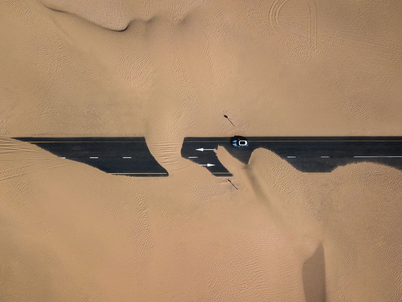 An aerial view of a road cutting across open desert, a single vehicle casting a long shadow over the sand