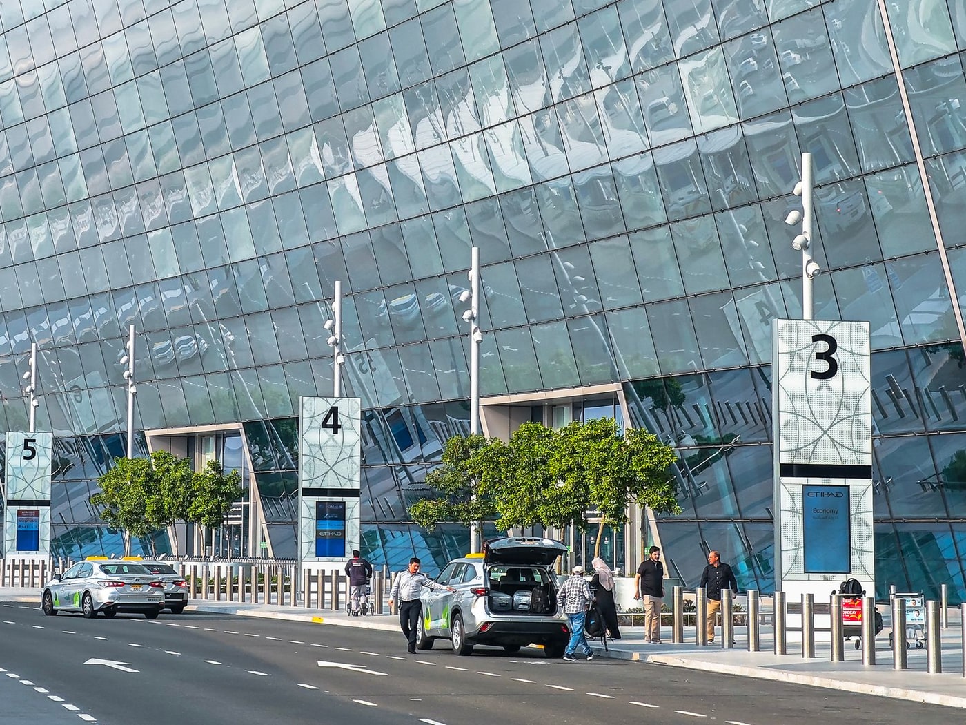 A terminal forecourt with numbered entrance gates, cars drawing up at the kerb and travellers crossing to the doors