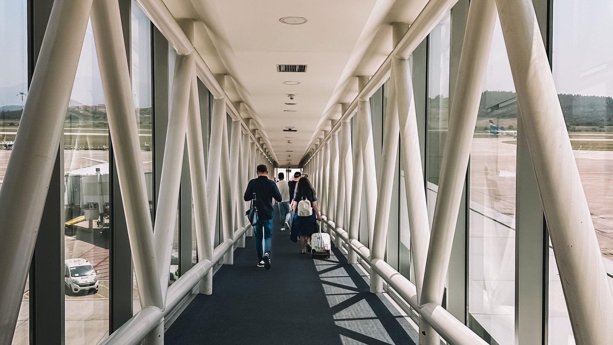 Passengers walking away down a white steel-and-glass jet bridge, the airport apron visible through the glass on both sides