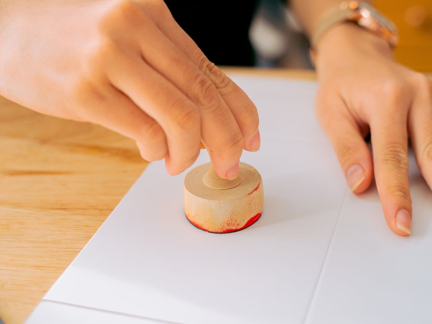 A wooden rubber stamp inked red being pressed onto a sheet of paper on a pale timber desk