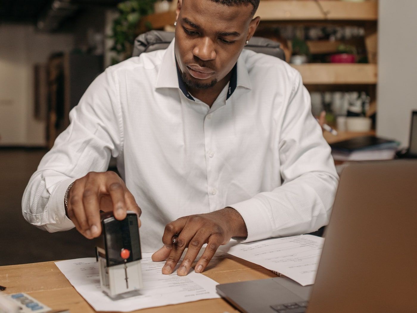 A man at a timber desk pressing a self-inking stamp onto a printed document, a laptop and further papers beside him